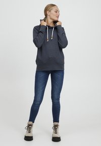 Hoodie gris foncé avec une capuche intérieure à motifs, des cordons blancs et des accents en bois. Associé à un jean skinny bleu et des bottes beiges.