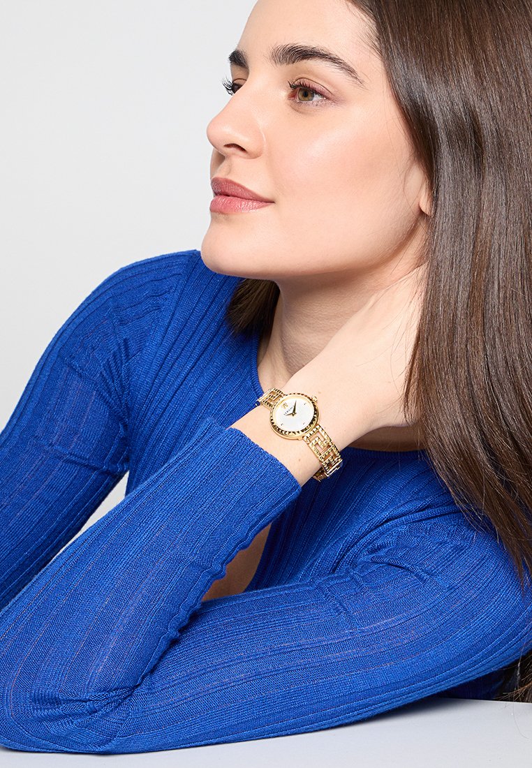 Montre-bracelet en métal doré avec un cadran blanc et des accents dorés. Portée sur un poignet, aux côtés d'un haut à manches longues bleu à côtes.