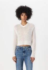 White, cropped knit sweater with a v-neck, ribbed cuffs, and hem. Paired with light blue jeans. Small logo on the chest.