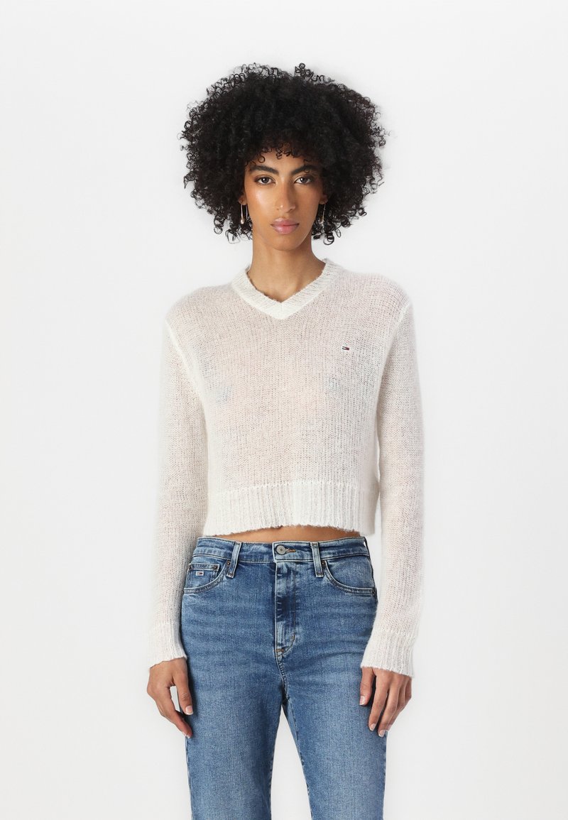 White, cropped knit sweater with a v-neck, ribbed cuffs, and hem. Paired with light blue jeans. Small logo on the chest.