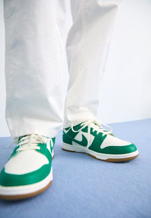 Białe spodnie i zielono-białe buty Nike z białymi sznurowadłami na jasnoniebieskiej podłodze na białym tle.