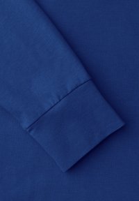 Rękaw niebieskiego, bawełnianego swetra o gładkiej fakturze, ściągaczu przy mankiecie i bezszwowej konstrukcji, bez widocznych wzorów czy elementów metalowych.