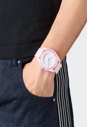 Rokas locītava ar rozā pulksteni, kas ielikta tumši zilās džinsu kabatā, uzvilkusi melnu krekli, rādot pulksteņa seju un siksniņas detaļas.