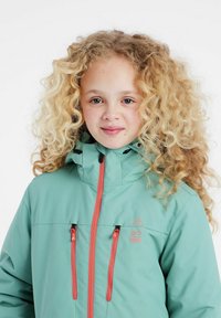 Veste de ski turquoise avec un col haut, deux poches avant zippées et des accents de fermeture éclair orange contrastants. Présente un logo sur le haut de la poitrine à gauche.