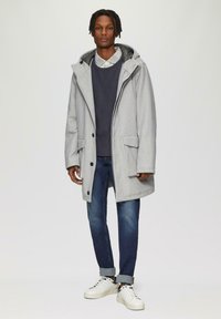 Grijze wollen buitenjas met een capuchon, twee voorkezen en zwarte knopen; gedragen over een marineblauwe trui, donkere jeans en witte sneakers.