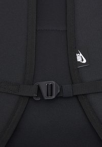 Zaino in nylon nero in primo piano che presenta un tessuto testurizzato, una tracolla regolabile e una fibbia di plastica. Etichetta con il logo Nike visibile.