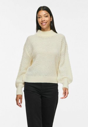 Jeune femme aux longs cheveux foncés portant un pull crème texturé et un pantalon noir, souriante sur fond blanc.