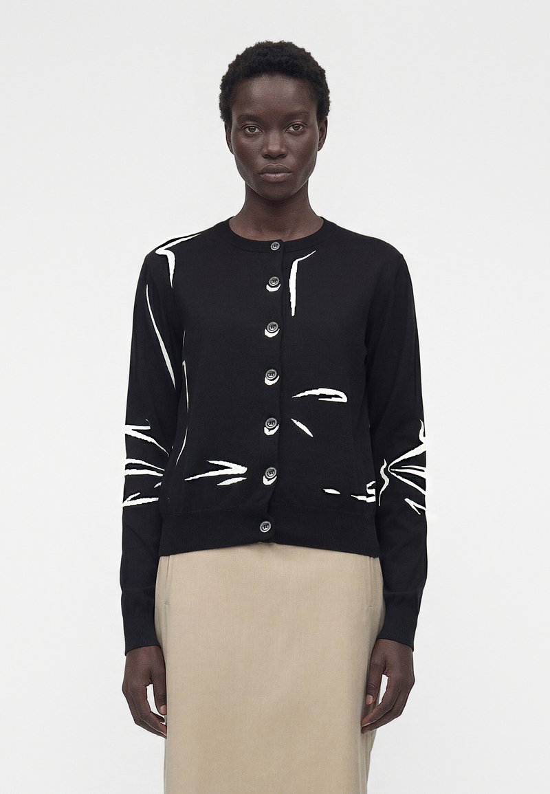 Γυναίκα που φοράει μαύρη ζακέτα με κουμπιά και λευκές αφηρημένες γραμμές και μπεζ φούστα, στέκεται μπροστά σε απλό φωτεινό φόντο.