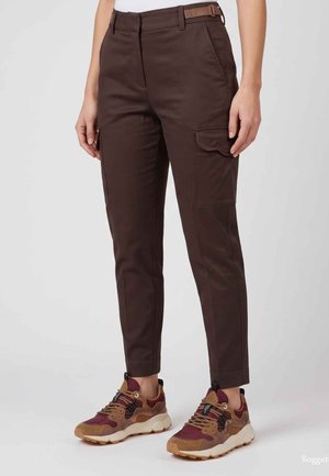 Persona che indossa pantaloni cargo slim marrone scuro con tasche a patta e sneakers marroni e bordeaux su sfondo chiaro.