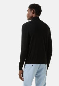 Černý pletený svetr s vysokým límcem, hladkou texturou, žebrovanými manžetami a lemem, v kombinaci s světle modrými kalhotami. Má jemné boční detaily.