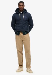 Felpa blu navy con tasca frontale e lacci bianchi, indossata sopra una camicia in tartan verde, abbinata a chinos beige e sneakers chiare.