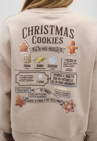 Felpa beige con una stampa della ricetta dei biscotti di Natale, accompagnata da icone degli ingredienti, tra cui il pan di zenzero, e dettagli decorativi.