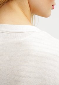 Ljusbeige stickad topp med vid halsringning, med en mjuk textur och subtil horisontell ribbing. Fokusera på området vid nacke bak.