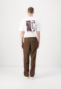 White t-shirt with a floral graphic in purple on the back, paired with brown trousers and white sneakers. Minimalist design with straight fit.