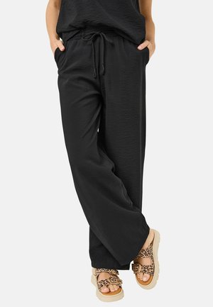 Zwarte losvallende broek met een gemêleerd oppervlak, voorzien van een elastische tailleband en een trekkoord. Geharmoniseerd met lichtgekleurde sandaal met luipaardprint.