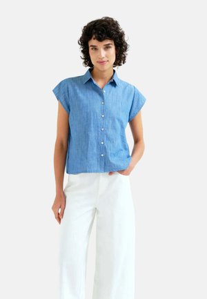 Mujer con cabello rizado y corto, que lleva una camisa azul sin mangas con botones y pantalones blancos de pierna ancha, de pie con una mano en el bolsillo.