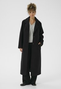 Woman with curly hair wearing a long black coat, gray buttoned cardigan, black pants, and black shoes standing against a white background.