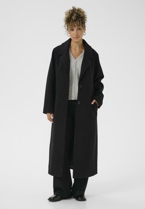 Woman with curly hair wearing a long black coat, gray buttoned cardigan, black pants, and black shoes standing against a white background.