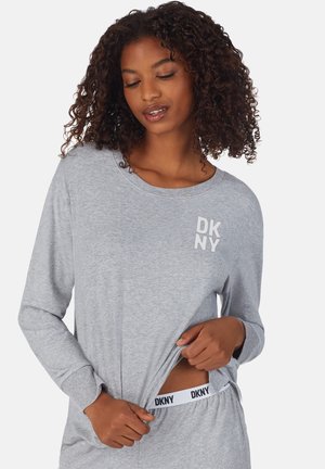 Frau mit lockigem Haar, die ein hellgraues langärmliges Oberteil und eine Hose trägt, zieht ihr Shirt hoch, um den weißen DKNY-Bund der Hose zu zeigen.