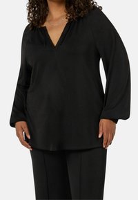 Svart långärmad topp med V-ringning och avslappnad passform, i slät tyg och minimalistisk design. Kombinerad med matchande byxor.