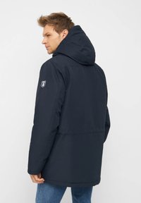 Giacca con cappuccio blu navy caratterizzata da una trama liscia, tasche frontali e un emblema sulla parte superiore della manica. Design semplice con vestibilità rilassata.