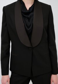 Musta bleiseri ruskealla satiinikauluksella, jossa on yksi napitus ja sivutaskut. Ominaisuuksiin kuuluu sileä pinta ja mittatilaustyönä tehty istuvuus.