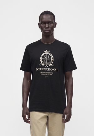 Άνδρας που φοράει μαύρο μπλουζάκι Nike International και μπεζ παντελόνια, στέκεται μπροστά σε λευκό φόντο, κοιτάζοντας μπροστά.