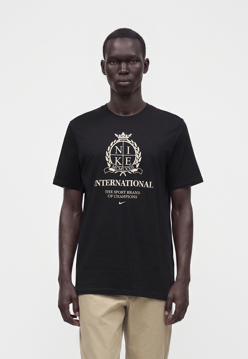 Άνδρας που φοράει μαύρο μπλουζάκι Nike International και μπεζ παντελόνια, στέκεται μπροστά σε λευκό φόντο, κοιτάζοντας μπροστά.