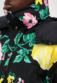 Chaqueta de plumas floral con base negra y patrones de flores en rosa, amarillo y verde. Cuello alto con detalles de botones y textura suave.