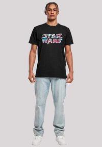 Černé bavlněné tričko s vícebarevným grafickým motivem "STAR WARS". Model nosí světle modré džíny a bílé tenisky.