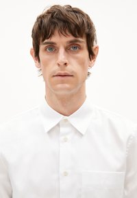 Junger Mann mit blauen Augen, kurzem braunem Haar, kleinen Creolen, der ein weißes Hemd mit Knöpfen vor einem schlichten hellen Hintergrund trägt.