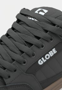 Black sneaker with a textured surface, flat laces, and white "GLOBE" logo. Features round eyelets and a contrasting brown sole.