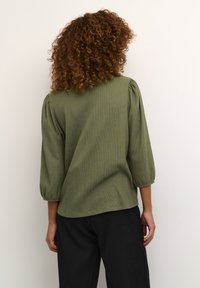 Femme aux cheveux bouclés portant un chemisier texturé vert olive à manches trois-quarts et un pantalon noir, debout contre un mur blanc uni.