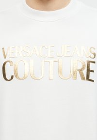 Λευκό ένδυμα με χρυσά μεταλλικά γράμματα "VERSACE JEANS COUTURE" στο ύψος του στήθους κάτω από στρογγυλή λαιμόκοψη.