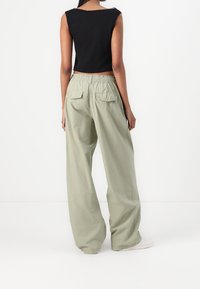 Persona vestida con pantalones cargo holgados de color verde claro con bolsillos traseros con solapa, una camiseta de tirantes negra y corta, y zapatillas blancas, de pie de espaldas.