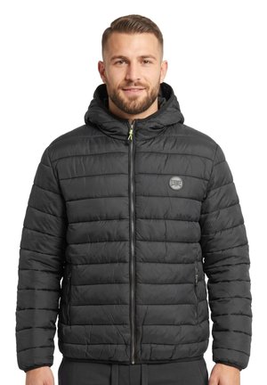 Un homme barbu porte une veste noire matelassée à capuche, avec une fermeture éclair sur le devant et un logo sur la poitrine, sur fond blanc.