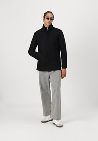 Svart, glidelåsjakke med ståkrage, med knapplukking, satt mot grå denim bukser og joggesko. Smooth tekstur, minimalistisk design.