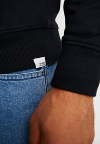 Sort sweatshirt med ribbede manchetter, båret over lyseblå denim. Liten hvid label forneden med "ISOLID est. 1999" trykt.