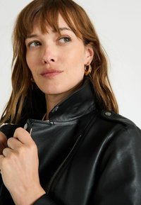 Femme aux cheveux châtain clair portant des boucles d'oreilles en or et une veste en cuir noir, tenant le col et regardant sur le côté.
