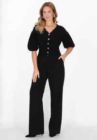 Schwarzes gestricktes Oberteil mit einem gewellten Ausschnitt und kurzen Puffärmeln, verziert mit dekorativen Knöpfen. Kombiniert mit hochgeschnittenen schwarzen Hosen.