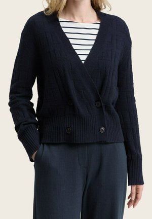 Mujer que lleva un suéter cruzado de color azul marino oscuro con patrones texturizados sobre una camisa blanca y negra a rayas y pantalones azul marino oscuro.