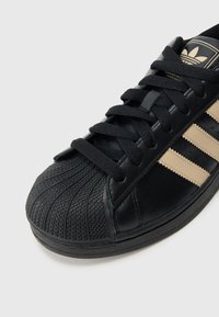 Detailní záběr na černou tenisku s béžovými pruhy, strukturovanou gumovou špičkou a černými tkaničkami na jednoduchém světlejším pozadí.