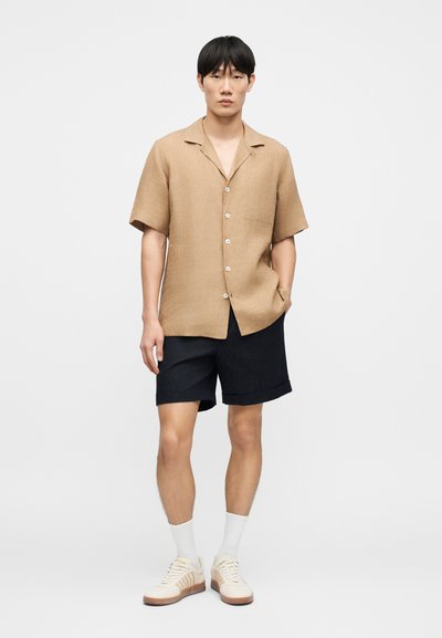 Μπεζ κοντομάνικο πουκάμισο με γιακά, κλείσιμο με κουμπιά και εμπρόσθια τσέπη, συνδυασμένο με σκούρα σορτς. Το μοντέλο φοράει λευκές κάλτσες και μπεζ παπούτσια.