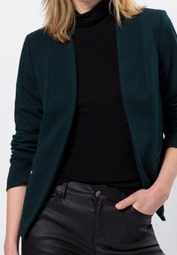 Blazer vert foncé à finition texturée, coupe slim, devant ouvert et revers crantés, associé à un col roulé noir et un pantalon noir.