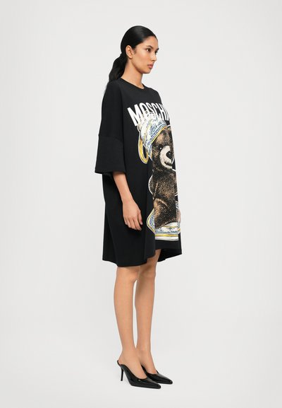 Melnā oversize T-kreklu kleita ar krāsainu lāča grafiku un tekstu "Moschino". Modelis valkā melnus augstos papēžus. Gluda tekstūra.