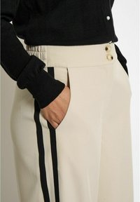 Beige trousers with black side stripes, featuring a relaxed fit, elastic waistband, and two decorative gold buttons at the top.