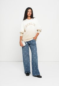 Sweatshirt bege e branco com o texto "BERLIM"; corte solto. Jeans de perna larga azul claro com padrão repetido de logotipo branco; sapatos pretos.