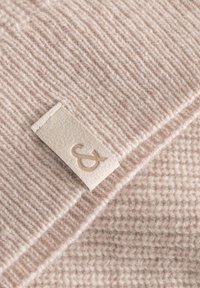 Beige Wollschal mit strukturiertem Rippenmuster und einem hellbraunen Etikett mit geprägtem Logo, das ein dezentes Designelement hinzufügt.