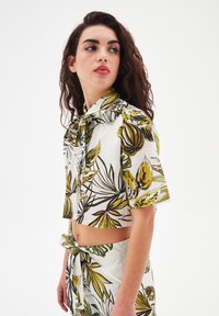 Blusa corta a maniche corte in cotone bianco con stampa botanica verde e gialla, dotata di colletto arricciato e chiusura con bottoni.