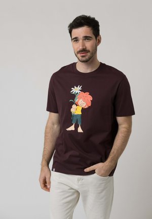 Mann mit kurzen dunklen Haaren und Bart, trägt ein dunkles T-Shirt mit einer Cartoonfigur, die eine große weiße Blume hält, weiße Hose, Hand in der Tasche.